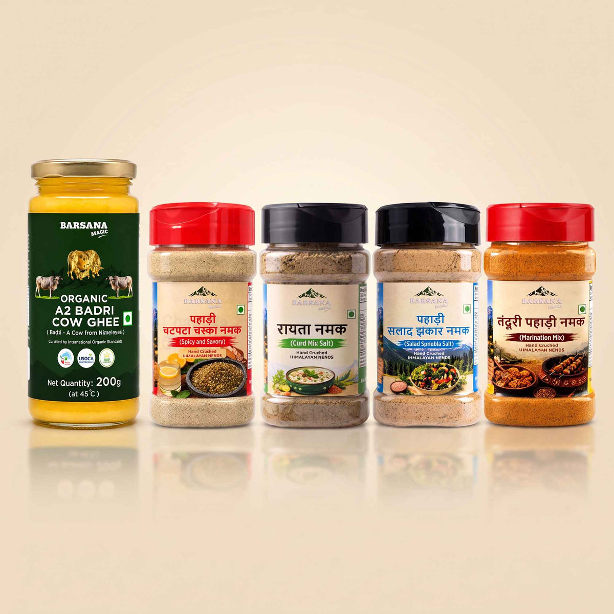 Barsana Magic and Five Indian spices and ghee products arranged in a row on a light beige background. From left to right: a large glass jar of Babsya Organic A2 Badri Cow Ghee with a gold lid and green label; a red-lidded jar of Panchachar Masala Chutney; a black-lidded jar of Chaashir Masala Chutney; a black-lidded jar of Panchachar Masala Chutney; and a red-lidded jar of Randhir Panchachar Masala Chutney.