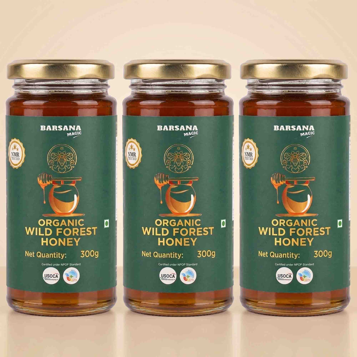 Barsana Magic and Three glass jars of Barsana Organic Wild Forest Honey with gold lids and dark green labels, arranged side by side on a light beige background.