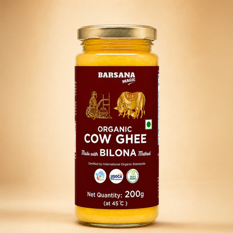 Barsana Magic andA 200g glass jar of Barsana Magic Organic Cow Ghee, made with Bilona Method, featuring a deep maroon label with gold illustrations and a gold metal lid, set against a warm beige background.