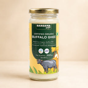 Barsana Magic and A 200g glass jar of Barsana Magic Certified Organic Buffalo Ghee, featuring a gold metal lid and a yellow and green label with a cow illustration, set against a warm beige background.