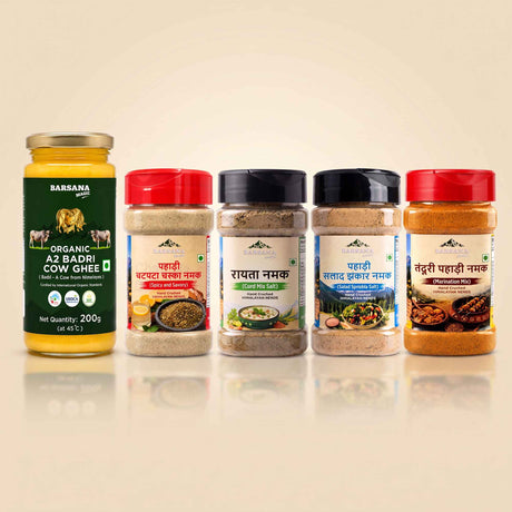 Barsana Magic and Five Indian spices and ghee products arranged in a row on a light beige background. From left to right: a large glass jar of Babsya Organic A2 Badri Cow Ghee with a gold lid and green label; a red-lidded jar of Panchachar Masala Chutney; a black-lidded jar of Chaashir Masala Chutney; a black-lidded jar of Panchachar Masala Chutney; and a red-lidded jar of Randhir Panchachar Masala Chutney.