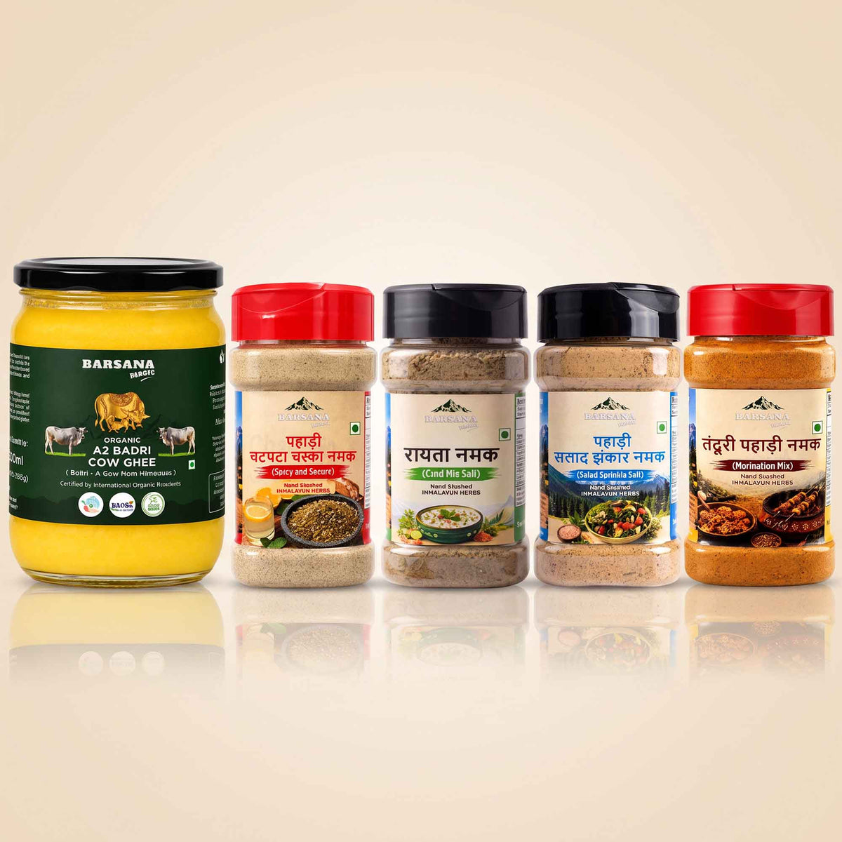 Barsana Magic and Five Barwana brand spices and ghee containers arranged in a row on a light beige background with a soft reflection below. The set includes a large jar of A2 Badhi Cow Ghee on the left, followed by three spice jars with red, white, and black lids, and a fourth spice jar with a red lid on the right.