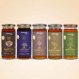 Barsana Magic and Five glass jars of BARSANA brand honey arranged in a row against a warm beige background. The jars have gold lids and labels in different colors: dark brown for Natural Ajwain Honey, purple for Natural Jamun Honey, brown for Natural Mustard Honey, olive green for Natural Acacia Honey, and dark green for Natural Neem Honey.