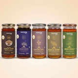 Barsana Magic and Five glass jars of BARSANA brand honey arranged in a row against a warm beige background. The jars have gold lids and labels in different colors: dark brown for Natural Ajwain Honey, purple for Natural Jamun Honey, brown for Natural Mustard Honey, olive green for Natural Acacia Honey, and dark green for Natural Neem Honey.