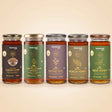 Barsana Magic and Five glass jars of BARANSA natural honey in different flavors and colors, lined up in a row against a warm beige background. The jars feature dark brown, green, purple, olive green, and dark green labels with gold lettering.