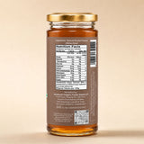 Barsana Magic and A glass jar of honey shown from the back, featuring a gold metal lid, a brown label with a visible Nutrition Facts panel, and a barcode. The honey inside is golden amber, and the background is a solid, warm beige.
