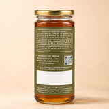 Barsana Magic and A glass jar of natural acacia honey with a gold metal lid and a dark olive green label, shown from the back against a solid beige background.