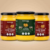 Barsana Magic and Three glass jars of Barsana Organic Cow Ghee arranged symmetrically on a warm beige background. The center jar has a dark green label, while the two side jars have dark red labels, all showing yellow ghee inside and black lids.