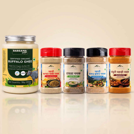 Barsana Magic and A group of Babsana brand Indian spices and ghee products arranged in a row on a reflective beige surface. The lineup includes a large jar of Certified Organic Buffalo Ghee on the left, followed by four spice jars with red, black, black, and red caps, showing various ground spices.