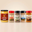 Barsana Magic and Five BARSANA brand Indian spices and ghee products arranged in a row on a light beige background. The lineup includes a large jar of Organic Cow Ghee on the left, followed by four spice jars with red, black, and dark blue lids and colorful labels.