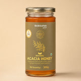 Barsana Magic and A 300g glass jar of Barsana Magic Natural Acacia Honey, featuring a metallic olive green label with gold lettering and a silver metal lid, set against a solid beige background.