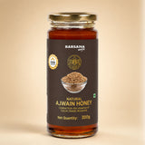 Barsana Magic and A 300g glass jar of Barsana Magic Natural Ajwain Honey, featuring a dark brown label with gold lettering and a gold metal lid, set against a solid beige background.