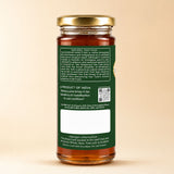 Barsana Magic and A glass jar of honey with a gold metal lid and a dark green label showing the back text and QR code, set against a solid beige background.