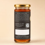 Barsana Magic and A glass jar of Natural A JWain Honey with a gold metal lid and a black label showing product details, set against a solid beige background.