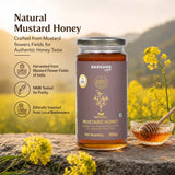 Barsana Magic and A jar of Barsana Natural Mustard Honey is displayed on a stone surface with a glass bowl of honey and a wooden dipper, set against a blurred background of yellow mustard flowers and hills. Text on the left highlights product features.