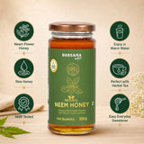 Barsana Magic and A 300g glass jar of Barsana Magic Natural Neem Honey, featuring a green label and a brown lid, displayed on a wooden surface with decorative honeycomb and green leaves. The jar is flanked by six circular green icons with white text highlighting product features like 'Neem Flower Honey', 'Raw Honey', and 'NMR Tested'.