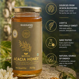 Barsana Magic and A 300g glass jar of Barsana Magic Natural Acacia Honey, featuring a gold cap and a brownish-gold label with gold text and illustrations. The jar is set on a stone surface with white flowers in the foreground, and a vertical column of marketing text and icons is displayed on the right against a blurred green background.