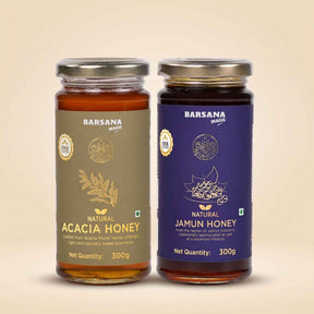 Barsana Magic and Two glass jars of Barsana Mashte honey on a light beige background. The left jar contains Acacia Honey with a brownish-olive label, and the right jar contains Jamun Honey with a dark purple label.