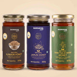 Barsana Magic and Three glass jars of Barsana Organic honey arranged side by side against a light beige background. The jars contain amber-colored honey and have distinct labels: dark brown for Awjain Honey, deep blue for Jamun Honey, and green for Van Tulsi Raw Honey.