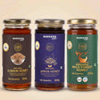 Barsana Magic and Three glass jars of Barsana Magic honey arranged side by side on a light beige background. The jars have gold lids and dark brown, deep purple, and green labels respectively, labeled Natural Ajwain Honey, Natural Jamun Honey, and Organic Wild Forest Honey.