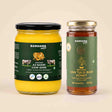 Barsana Magic and Two glass jars of Barsana brand products on a light beige background: a jar of Az Badri Cow Ghee with a black lid and yellow contents, and a jar of Van Tulsi Raw Honey with a gold lid and green label.