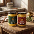 Barsana Magic and Two jars of Barsana organic cow ghee displayed on a rustic wooden table in a warm, traditional kitchen setting. One jar is labeled 'A2 Badhi Cow Ghee' with a dark green label, and the other is 'Organic Cow Ghee' with a dark red label.