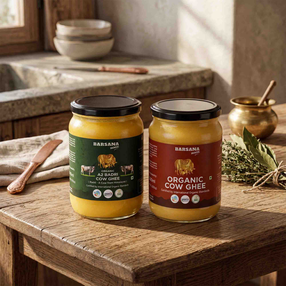 Barsana Magic and Two jars of Barsana organic cow ghee displayed on a rustic wooden table in a warm, traditional kitchen setting. One jar is labeled 'A2 Badhi Cow Ghee' with a dark green label, and the other is 'Organic Cow Ghee' with a dark red label.