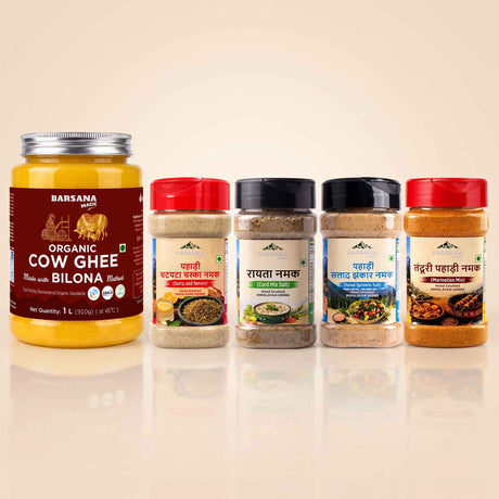 Barsana Magic and Four BarSana Namak spice products arranged in a row on a light beige background: a large jar of organic cow ghee bilona with a red label, and three smaller spice jars with red, black, and brown lids and beige, gray, and brown contents.