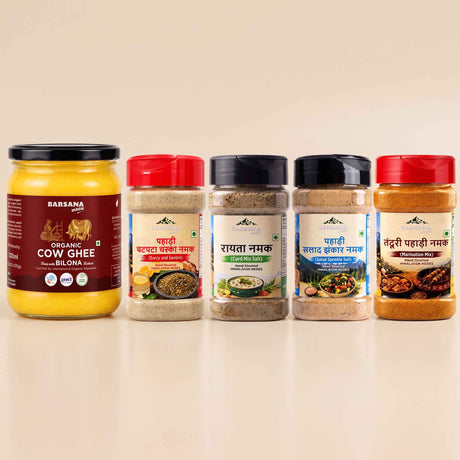Barsana Magic and Five spice and ghee jars arranged in a row on a light beige background. The leftmost jar is a large glass jar of Barsana Organic Cow Ghee with a yellow contents and dark red label.
