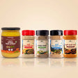 Barsana Magic and Five jars of Barana Brand spices and dairy products arranged in a row on a light beige background with a soft reflection below. The products include a large jar of Herbal Dairy Spread on the left, followed by four spice jars with red, black, black, and red caps.