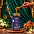 Barsana Magic and A jar of Barsana Jamun Honey is shown on a wooden coaster with honey dripping from a wooden dipper. A hand with henna is visible holding the dipper.