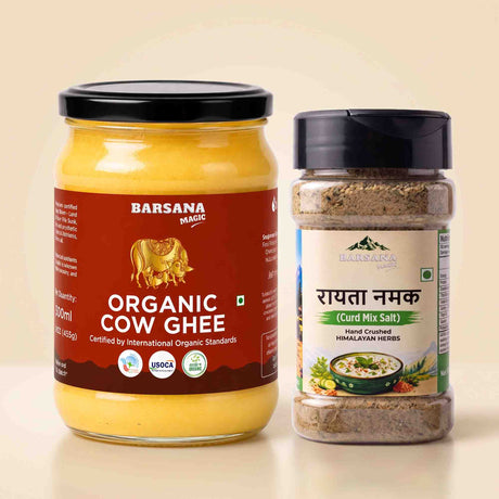 Barsana Magic and A glass jar of Barsana Magic Organic Cow Ghee with a black lid and a brown label, next to a clear plastic jar of Barsana Magic Rayyata Namak (Curd Mix Salt) with a black lid and beige label, set against a light beige background.