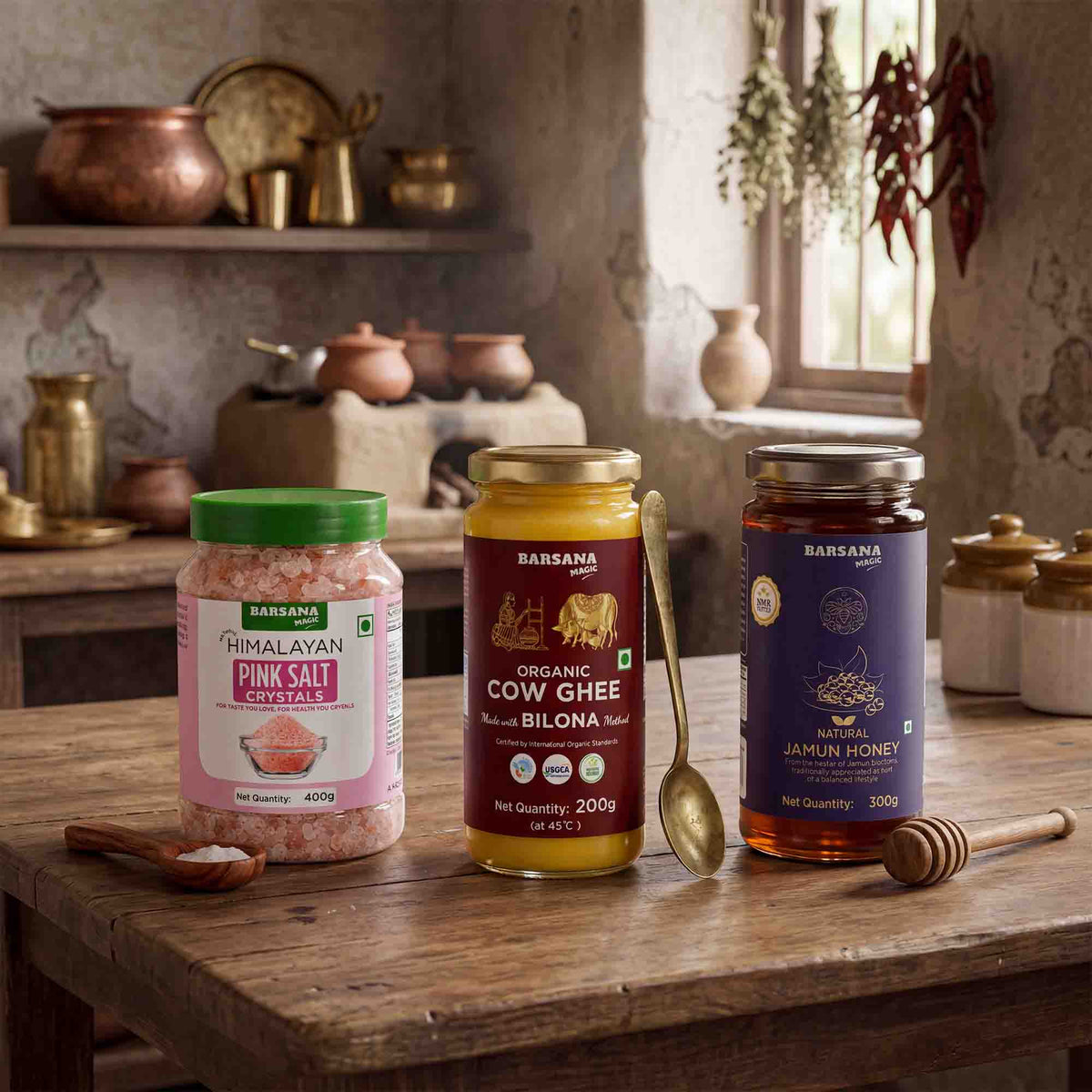 Barsana Magic and Three jars of Barsana natural products arranged on a rustic wooden table in a warm, vintage-style kitchen setting: a jar of Himalayan Pink Salt, a jar of Organic Cow Ghee, and a jar of Jamun Honey, with spoons and honey dipper nearby.
