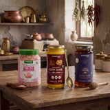 Barsana Magic and Three jars of Barsana natural products arranged on a rustic wooden table in a warm, vintage-style kitchen setting: a jar of Himalayan Pink Salt, a jar of Organic Cow Ghee, and a jar of Jamun Honey, with spoons and honey dipper nearby.