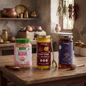 Barsana Magic and Three jars of Barsana natural products arranged on a rustic wooden table in a warm, vintage-style kitchen setting: a jar of Himalayan Pink Salt, a jar of Organic Cow Ghee, and a jar of Jamun Honey, with spoons and honey dipper nearby.