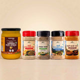 Barsana Magic and Five packaged food products arranged on a beige surface against a warm beige background: a large jar of BARSANA Turmeric Dairy Spread on the left, followed by four smaller mountain-style spice or seasoning jars with red, white, black, and red lids. No people or real environment are present, so this is Product/Packaging.