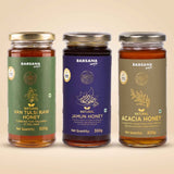Barsana Magic and Three glass jars of Barsana Magic natural honey lined up against a solid beige background. From left to right, the labels are green for Van Tulsi Raw Honey, dark purple for Jamun Honey, and olive green for Acacia Honey.