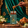 Barsana Magic and A jar of Barsana Organic Wild Forest Honey is shown on a wooden surface with honey dripping from a wooden dipper into the jar. A hand with henna is visible holding the dipper.