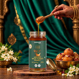 Barsana Magic and A jar of Barsana Organic Wild Forest Honey is shown on a wooden surface with honey dripping from a wooden dipper into the jar. A hand with henna is visible holding the dipper.