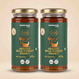 Barsana Magic and Two glass jars of Barsana Handmade Organic Wild Forest Honey with gold lids and dark green labels, displayed side-by-side on a light beige background.